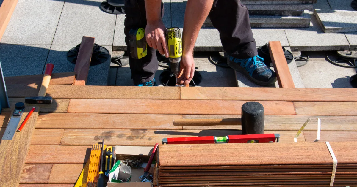 a deck builder building a wooden deck, using a power drill on teak or similar hardwood planks