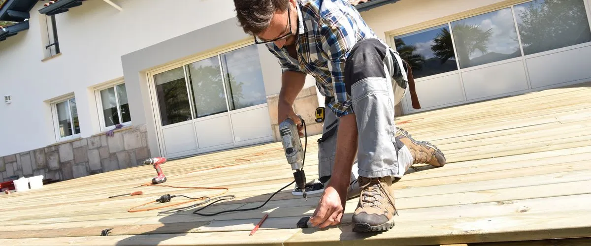 A contractor kneeling and using a power drill to secure wooden planks while performing an outdoor deck repair in front of a home with sliding glass doors.