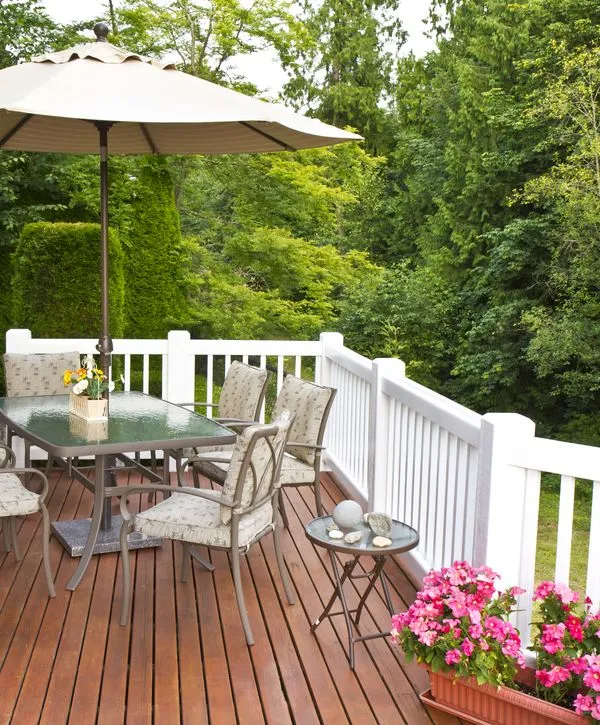 A beautiful backyard wooden deck installation in Nashville, TN, featuring classic white railings, a glass-top dining set with an umbrella, and bright pink potted petunias surrounded by lush green trees.