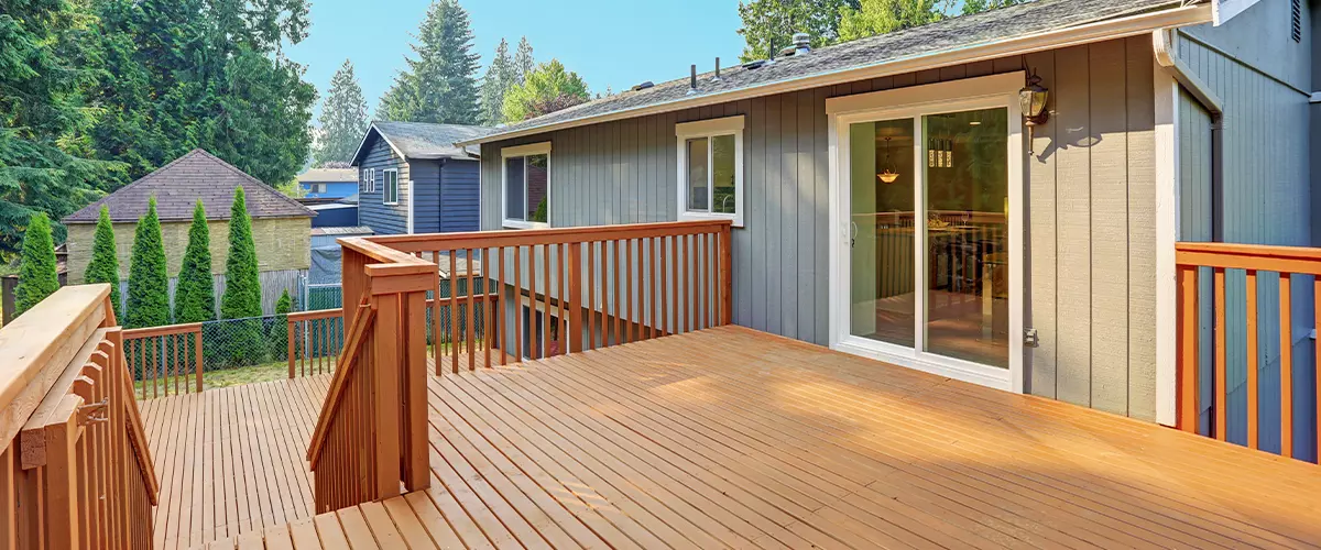An elevated backyard wooden deck with matching wood railings and stairs, connected to the sliding glass door of a gray residential home.
