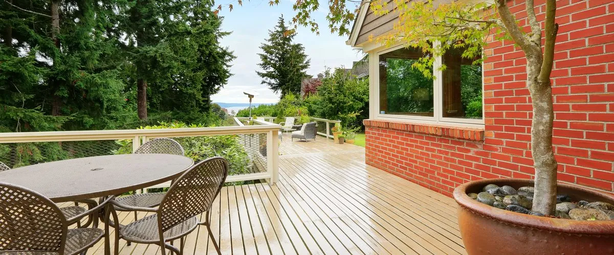 A light-colored outdoor deck attached to a red brick house, equipped with a round metal mesh dining set, wire railings, and a large potted tree with lush backyard scenery in the background.
