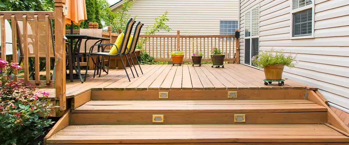 A low-level backyard wooden deck featuring wide illuminated stairs, a patio dining set with yellow pillows, and potted plants along the railing.