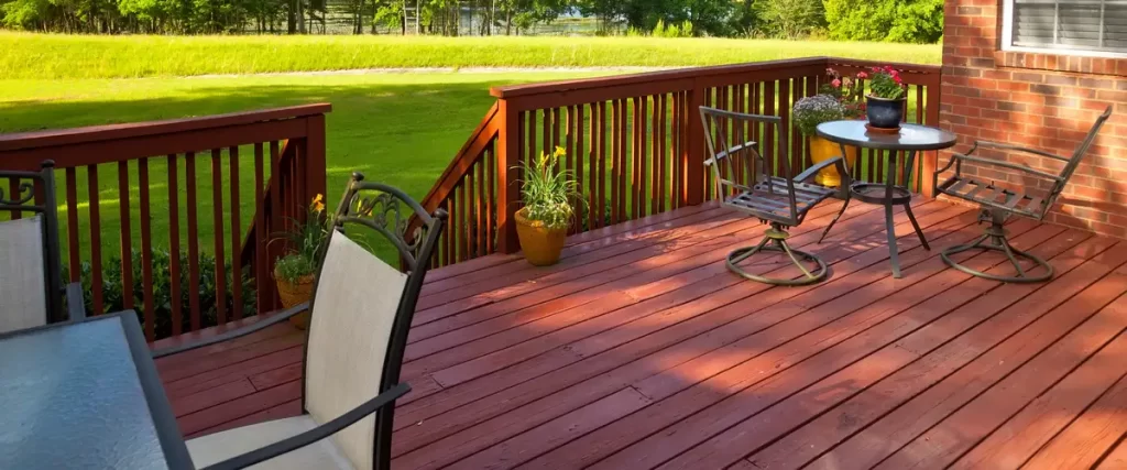 A reddish-brown stained wood deck equipped with a glass-top patio table, metal chairs, and vibrant potted plants overlooking a large grassy backyard.