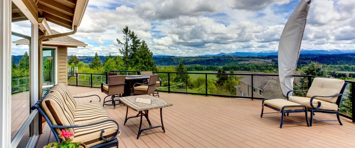 A spacious outdoor deck featuring modern glass and metal railings, a fire pit table, and cozy lounge furniture with striped cushions overlooking a scenic valley and mountain landscape.