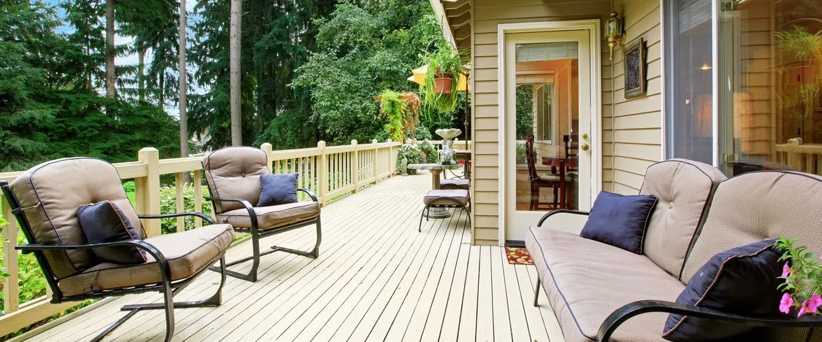 A spacious outdoor deck attached to a beige home, featuring light-colored decking and comfortable metal patio sofas with tan cushions and navy blue pillows.