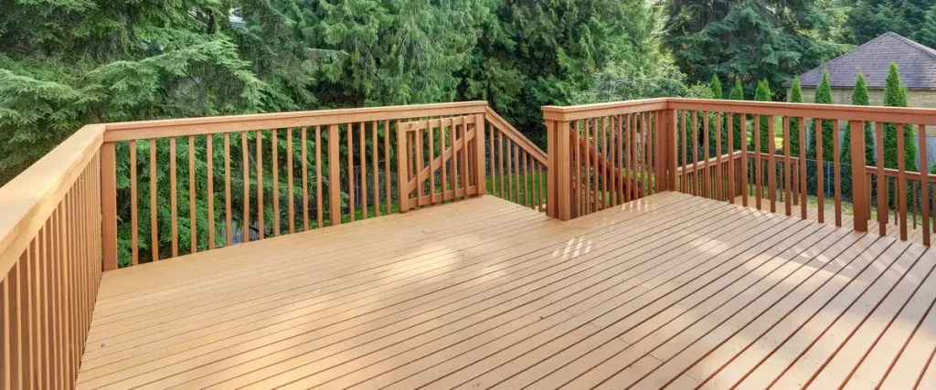 A spacious, empty wooden deck featuring matching wood railings and a small safety gate at the top of the stairs, surrounded by dense green trees.