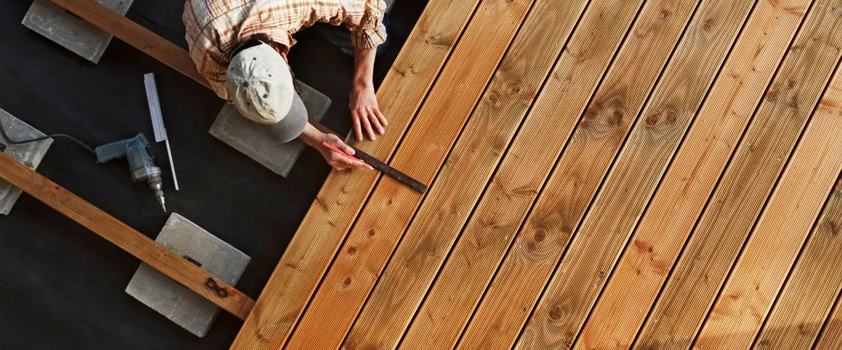 Top-down view of a worker using a square ruler and pencil to measure and mark wooden planks before cutting during a deck repair project.